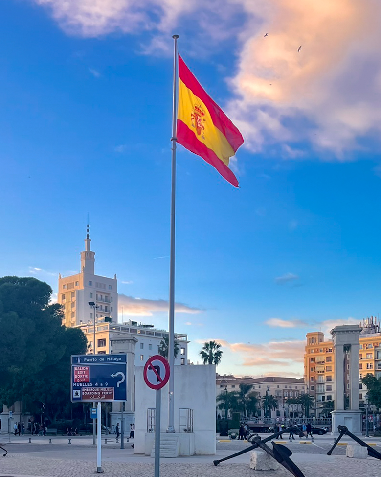 Bandera española con el emblema de la Autoridad Portuaria de Málaga en Muelle Tres, con el Edificio de Equitación al fondo