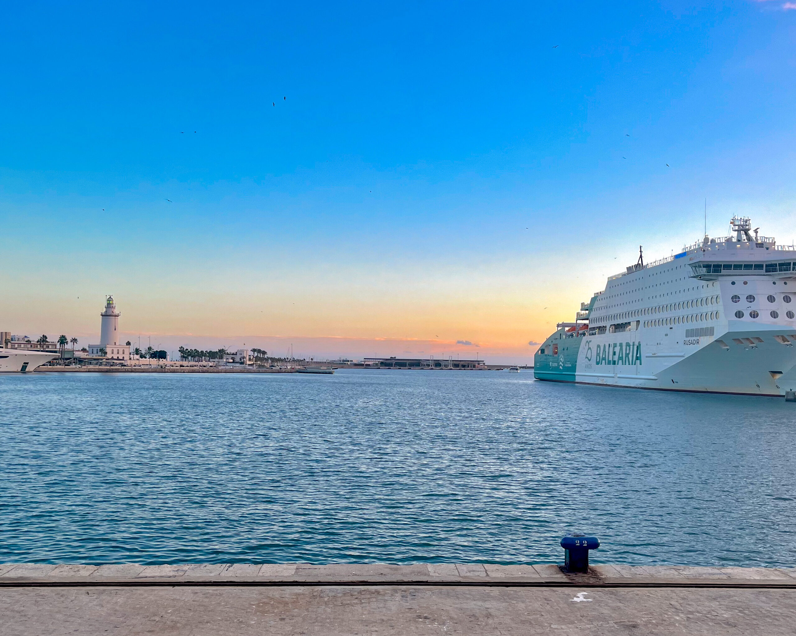 Hafen von Málaga mit einem Balearia-Kreuzfahrtschiff, gesehen vom Muelle Dos, mit dem Leuchtturm La Farola im Hintergrund bei Sonnenuntergang