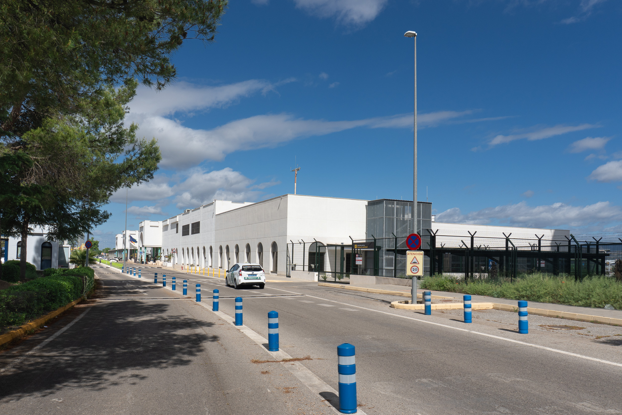 Federico García Lorca Airport Granada-Jaén, vía de acceso y edificio terminal, con bolardos azules, pinos y un cielo despejado